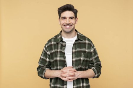 Portrait photo of a cheerful, bearded man smiling joyfully with intertwined fingers, wearing a plaid shirt, posed against an isolated yellow background, looking at the camera