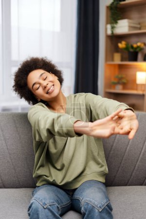 Téléchargez les photos : Femme afro-américaine joyeux étirement, se réveiller les yeux fermés assis sur un canapé confortable à la maison. Concept du matin, détente - en image libre de droit