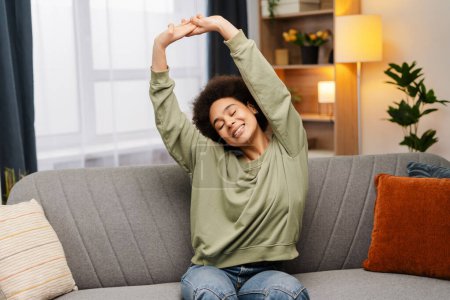 Photo for Portrait of smiling attractive African American woman stretching, wake up with closed eyes sitting on comfortable sofa at home. Morning concept, relaxing - Royalty Free Image