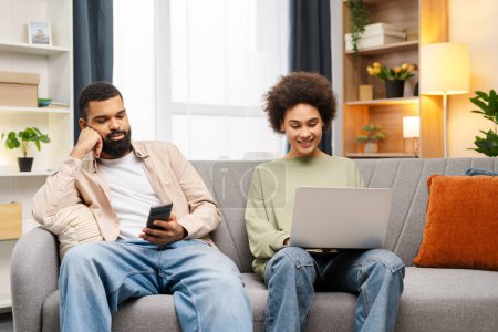 Foto de Hermosos freelancers afroamericanos usando un ordenador portátil, sosteniendo el teléfono móvil, comprando en línea mientras están sentados en casa en un cómodo sofá. Atractivo proyecto de planificación de compañeros de trabajo jóvenes. Concepto de trabajo en equipo - Imagen libre de derechos