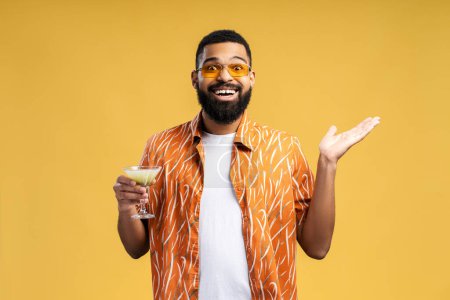 Téléchargez les photos : Un bel homme afro-américain excité portant des lunettes de soleil tenant un cocktail, regardant la caméra. Élégant hipster debout isolé sur fond jaune. Concept de fête, vacances, célébration - en image libre de droit