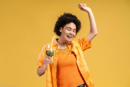 Téléchargez les photos : Femme afro-américaine excitée et souriante tenant un cocktail avec les yeux fermés dansant, s'amusant isolé sur fond jaune. Belle femme portant une chemise orange. Concept d'été, célébration de fête - en image libre de droit
