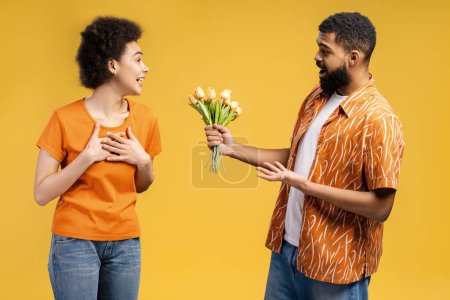 Foto de Atractiva pareja afroamericana, joven barbudo dando tulipanes amarillos a mujer excitada con el pelo rizado, de pie aislado sobre fondo amarillo. Concepto de celebración, cumpleaños - Imagen libre de derechos