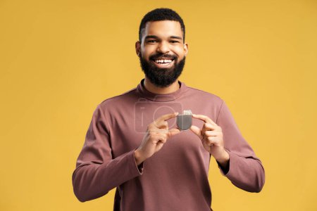 Photo for Smiling African American man holding pacemaker, cardioverter defibrillator looking at camera. Attractive patient standing isolated on yellow background. Health care concept, cardio treatment - Royalty Free Image
