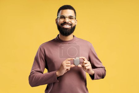 Smiling, attractive African American man holding pacemaker looking at camera. Patient showing cardioverter defibrillator standing isolated on yellow background. Concept of health care, support