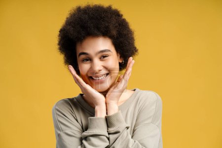 Foto de Retrato de una atractiva mujer afroamericana sonriente con el pelo rizado mirando a la cámara aislada sobre fondo amarillo. Concepto de cuidado de la piel, belleza natural - Imagen libre de derechos