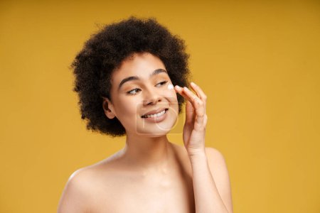 Foto de Hermosa mujer afroamericana sonriente con el pelo rizado aplicando crema en la cara mirando hacia otro lado de pie aislado sobre fondo amarillo. Concepto de belleza, cuidado de la piel, cosmetología - Imagen libre de derechos