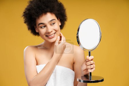 Téléchargez les photos : Jolie, heureuse femme afro-américaine en serviette blanche regardant dans le miroir après la douche, routine du matin. Modèle de mode souriant avec cheveux bouclés sur fond jaune. Concept d'hygiène, soin de la peau - en image libre de droit