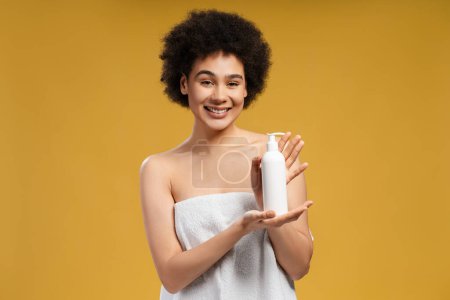 Photo for Beautiful African American young woman with curly hair in white towel after shower holding white bottle, mockup looking at camera. Advertisement concept, hygiene - Royalty Free Image