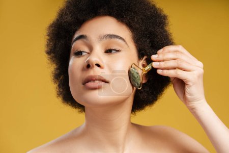 Photo for Attractive African American woman with curly hair doing face massage with massage roller, looking away, standing isolated on yellow background. Concept of skin care, spa, anti aging - Royalty Free Image