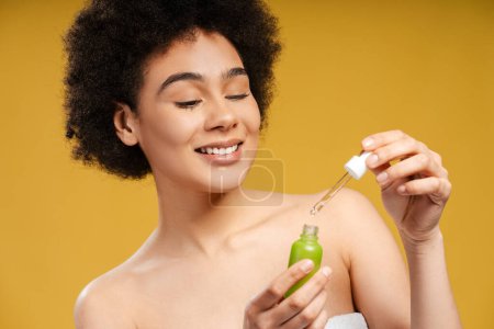 Foto de Hermosa joven latina sosteniendo botella verde aplicando suero a la cara, de pie aislada sobre fondo amarillo. Concepto de cuidado de la piel, procedimiento cosmético, hidratante - Imagen libre de derechos