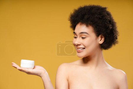 Foto de Atractiva mujer latina sonriente con el pelo rizado sosteniendo tarro blanco de crema hidratante, mirando hacia otro lado de pie aislado sobre fondo amarillo. Cosmetología conceptual, producto cosmético - Imagen libre de derechos
