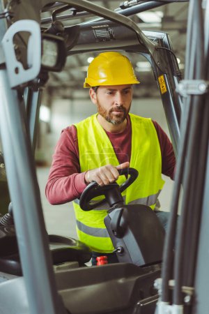 Photo for Attractive bearded man wearing yellow protective helmet and workwear driving forklift, working in warehouse. Concept of transportation, vehicle - Royalty Free Image