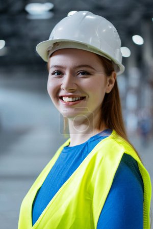 Téléchargez les photos : Portrait de belle, souriante, jeune femme portant un casque et des vêtements de travail, regardant la caméra. Travailleur professionnel travaillant dans un entrepôt. Concept industriel, entreprise - en image libre de droit