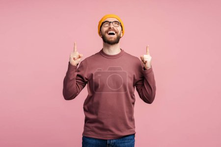 Photo for Amazed middle aged caucasian bearded man 40s, in eyeglasses and yellow hat pointing his index fingers up, expressing WOW emotion, amazement isolated on blue background - Royalty Free Image