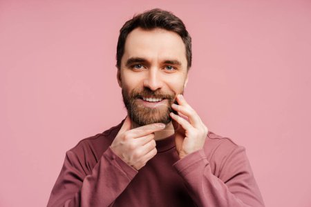Photo for Closeup portrait of handsome smiling man touching his face, correct beard looking at mirror isolated on pink background. Skin care, hygiene, morning routine concept - Royalty Free Image