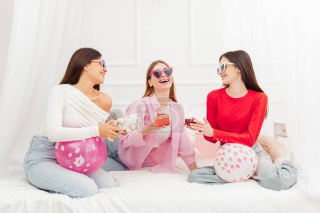 Photo for Group of overjoyed beautiful girls, best friends wearing eyeglasses communicating, celebration birthday, having fun with balloons sitting on bed. Holiday concept - Royalty Free Image