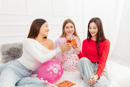 Foto de Grupo de mujeres hermosas sonrientes tostando vino, comiendo dulces de chocolate, durante la celebración despedida de soltera sentada en el sofá de casa. Concepto de cumpleaños - Imagen libre de derechos