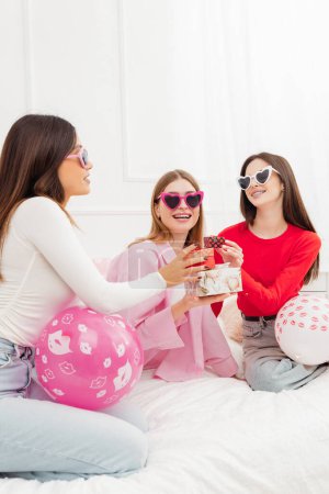 Photo for Vertical view of the overjoyed beautiful girls, best friends wearing sunglasses holding gifts and celebrating birthday, having fun with balloons sitting on bed. Holiday concept - Royalty Free Image