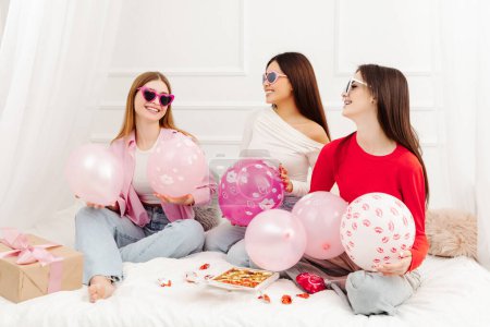 Foto de Feliz hermosas mujeres multirraciales con gafas de sol elegantes sentados en la cama mirándose. Jóvenes modelos de moda atractiva posando para fotos - Imagen libre de derechos