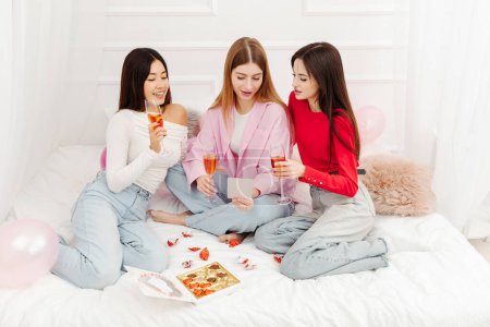 Foto de Grupo de sonrientes mujeres hermosas multirraciales bebiendo vino, comiendo dulces de chocolate, hablando celebración cumpleaños sentado en casa. concepto de despedida de soltera - Imagen libre de derechos