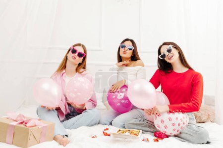 Foto de Feliz hermosas mujeres multirraciales con gafas de sol elegantes sentados en la cama y mirando a la cámara. Jóvenes modelos de moda atractiva posando para fotos - Imagen libre de derechos