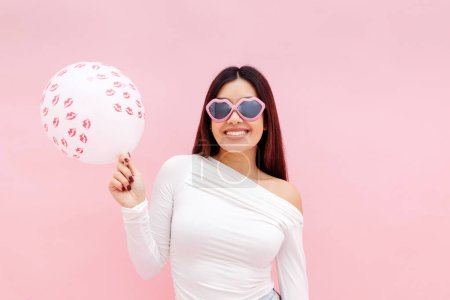 Photo for Cheerful brunette asian lady wearing sunglasses holding balloon and looking at camera while posing isolated on pink background. Valentines day celebration concept - Royalty Free Image
