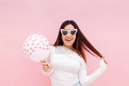 Foto de Mujer alegre excitada en gafas de sol posando a cámara con lindo globo aislado sobre fondo rosa. Concepto de vacaciones - Imagen libre de derechos