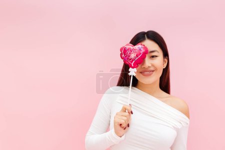 Téléchargez les photos : Heureuse dame asiatique souriante couvrant l'œil avec un ballon et regardant la caméra tout en posant isolé sur fond rose. Concept de célébration de la Saint-Valentin - en image libre de droit