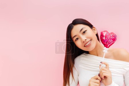 Téléchargez les photos : Portrait de dame asiatique brune joyeuse tenant ballon et regardant la caméra tout en posant isolé sur fond rose. Concept de célébration de la Saint-Valentin - en image libre de droit