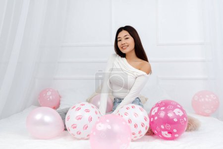 Foto de Linda morena mujer asiática sentada alrededor de globos rosados y mirando a la cámara mientras está sentada en un acogedor apartamento y celebrando su cumpleaños - Imagen libre de derechos