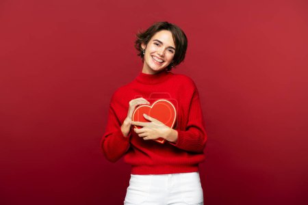 Foto de Hermosa mujer joven sonríe ante la cámara, mientras sostiene una caja de regalo roja, aislada sobre fondo rojo. Un lindo regalo para San Valentín o cualquier otro evento - Imagen libre de derechos