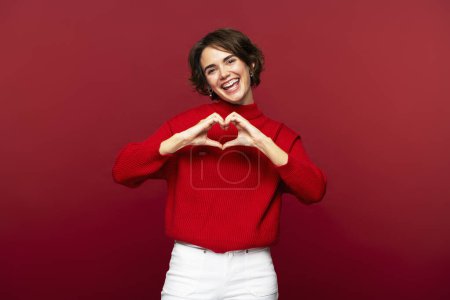 Portrait photo of joyful, attractive woman making a heart gesture with her hands, celebrating Valentine's Day, posing isolated on a red background, and looking into the camera 