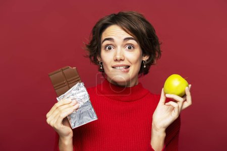 Téléchargez les photos : Femme réfléchie, tenant soigneusement une barre de chocolat et une pomme, choisissant entre une nourriture saine et malsaine, isolée sur une toile de fond rouge, regardant la caméra. Concept de régime alimentaire - en image libre de droit