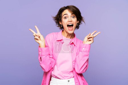 Foto de Emocionada hermosa mujer con elegante camisa rosa que muestra signo de victoria y mirando a la cámara con la boca abierta aislada sobre fondo violeta. Concepto de publicidad - Imagen libre de derechos