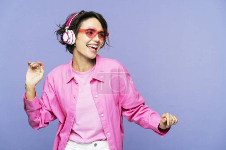 Foto de Hermosa mujer sonriente en gafas de sol con auriculares escuchando música, mirando hacia otro lado aislada sobre fondo violeta, bailando, divirtiéndose. concepto de estilo de vida feliz - Imagen libre de derechos