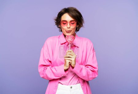 Foto de Mujer linda sonriente con ropa rosa y gafas de sol, sosteniendo el pop lolly, mostrando su lengua, de pie aislado sobre fondo violeta. Concepto de postres, comida - Imagen libre de derechos