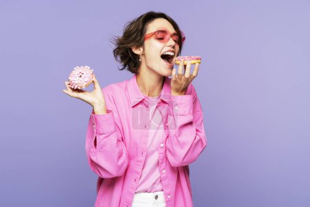 Foto de Retrato de mujer excitada sosteniendo rosquillas rosadas, postre mordedor aislado sobre fondo violeta. Concepto de comida, postre - Imagen libre de derechos