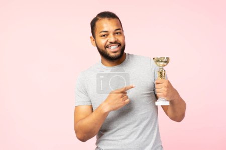 Foto de Overjoy sonriente hombre afroamericano vistiendo camiseta casual gris sosteniendo copa de trofeo aislado sobre fondo rosa. Varón positivo usando aparatos ortopédicos mirando la cámara. Concepto de victoria - Imagen libre de derechos