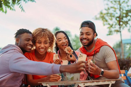 Photo for Group of smiling stylish African American friends taking selfie, eating pizza, drinking lemonade sitting together in modern cafe - Royalty Free Image