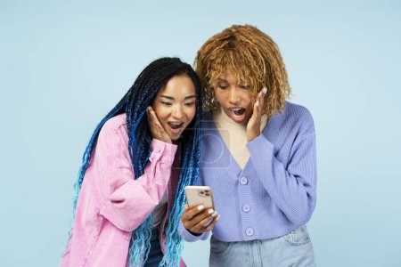 Photo for Portrait of overjoyed women friends holding mobile phone shopping online with sales, sport betting, celebration success isolated on blue background - Royalty Free Image