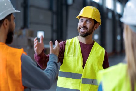 Photo for Handsome, smiling, bearded, middle aged man wearing protective helmet shaking hands with colleagues standing in warehouse, greetings, deal. Concept of cooperation, communication - Royalty Free Image