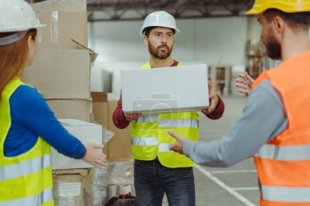 Photo for Handsome middle aged worker wearing hard hat and uniform, holding box, talking with colleagues in warehouse. Concept of teamwork, cooperation - Royalty Free Image