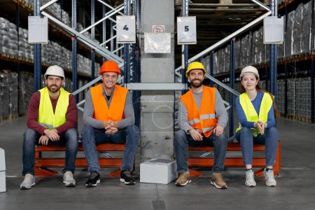 Téléchargez les photos : Travailleurs confiants, heureux, souriants, employés portant un casque et vêtements de travail assis dans l'entrepôt, regardant la caméra, réunion. Concept de travail d'équipe - en image libre de droit