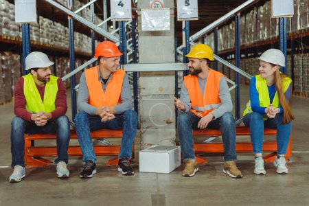 Téléchargez les photos : Des travailleurs professionnels, des ingénieurs qui portent des chapeaux durs et des vêtements de travail qui parlent, discutent de quelque chose, sont assis dans un entrepôt. Concept d'industrie, logistique - en image libre de droit