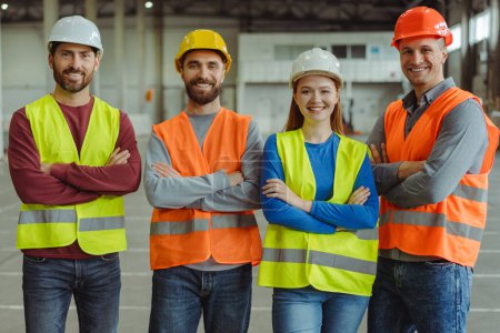 Photo for Smiling confident workers, engineers wearing hard hats and work wear with arms crossed looking at camera, posing in warehouse. Concept of teamwork, logistics, industry - Royalty Free Image
