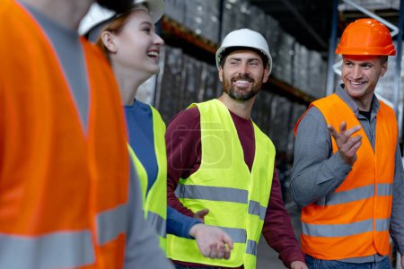 Photo for Group of smiling managers wearing hard hats and vests, walking through large warehouse, discussing cooperation, distribution, planning. Teamwork concept - Royalty Free Image