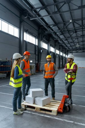 Photo for Group of successful warehouse managers wearing hard hats and vests loading boxes pallet trucks, talking while working in warehouse. Concept of industry, logistics, storage - Royalty Free Image