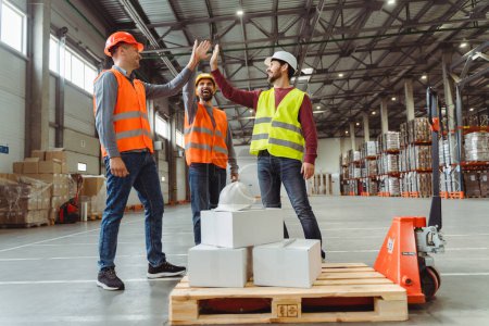Photo for Happy, confident workers, managers wear hard hats, vests, work wear giving high fives, work together in warehouse, standing near pallet of trucks with boxes. Concept of successful teamwork - Royalty Free Image