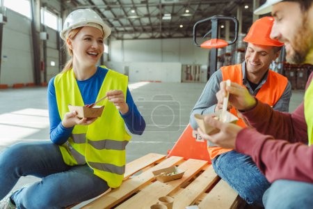 Photo for Happy, smiling workers, factory managers wearing hard hats, vests eating meal, sitting, talking while having lunch together in warehouse. Food concept - Royalty Free Image